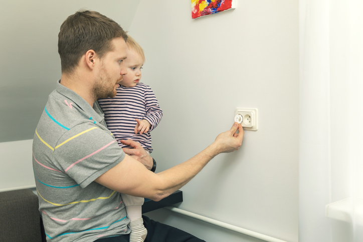 Tētis tur rokās mazu bērnu un aizver kontaktdakšu ar elektrības kontaktdakšu.